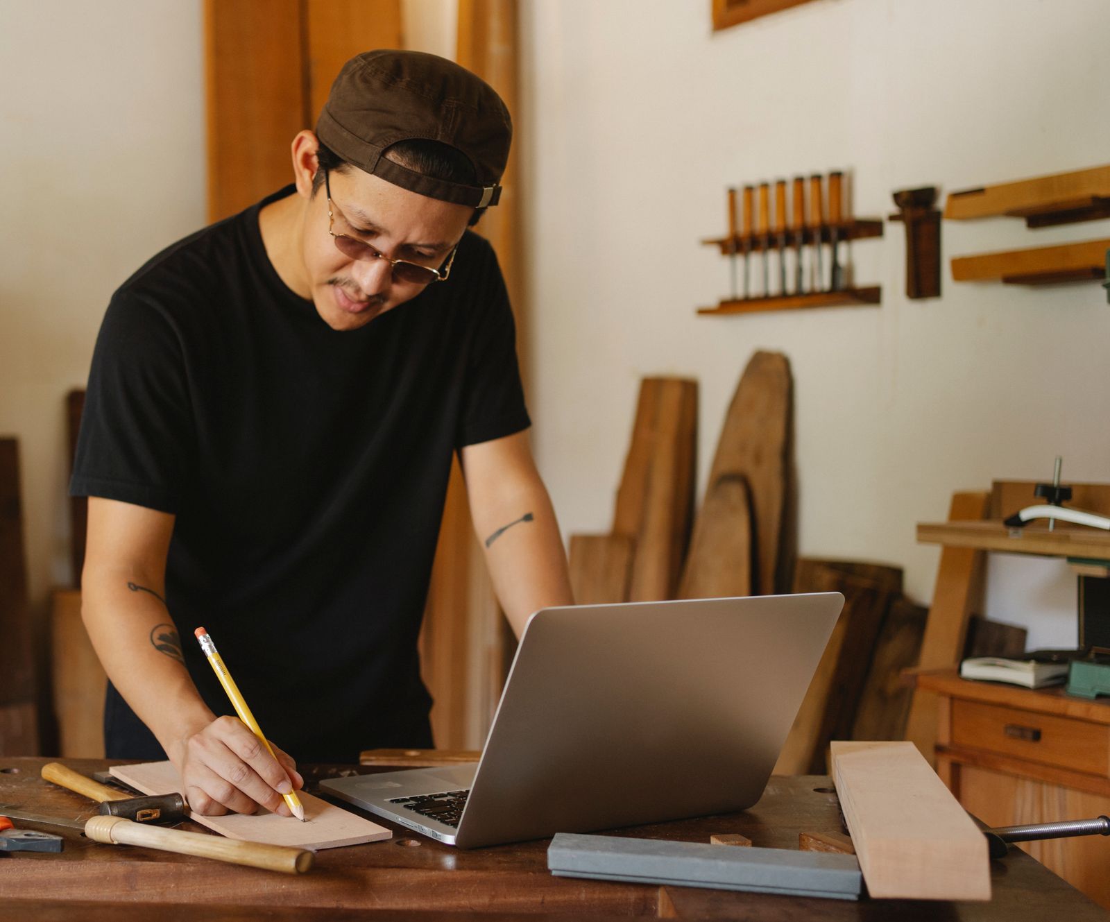 A carpenter taking notes beside a laptop at a workbench in a woodworking shop.