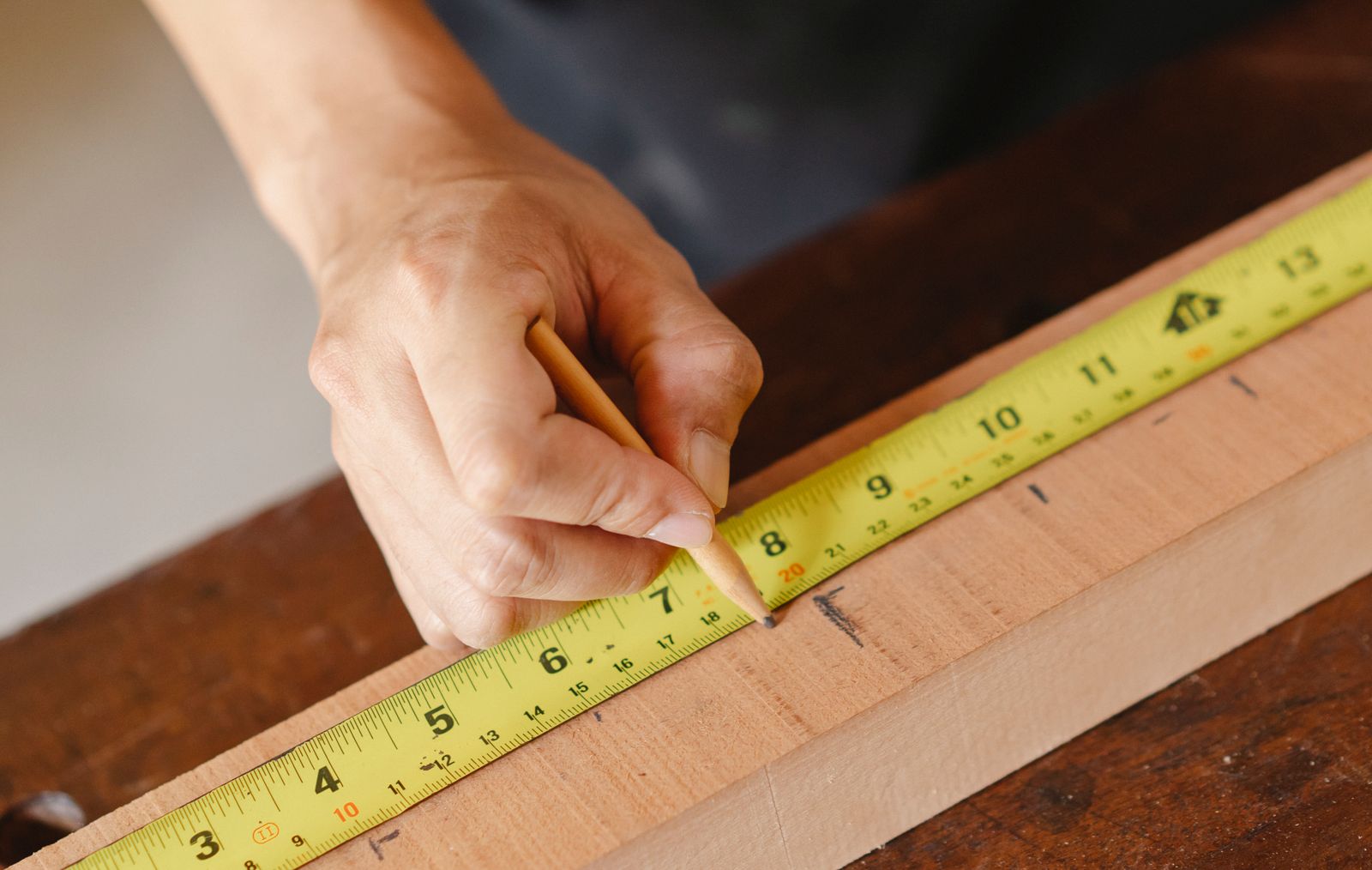 A hand marking a wooden plank with a pencil and measuring tape.